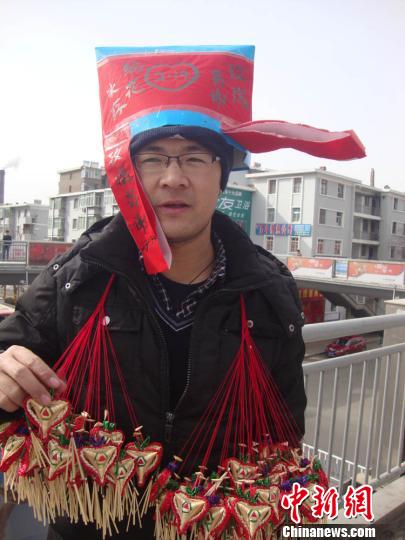 西北师大大四学生祁一桐头戴自己制作的“僧帽”，帽子上写着“鲜花易谢，编花长存”，在甘肃白银街头卖编花。