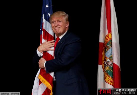 当地时间2016年10月24日，美国佛罗里达州坦帕市，美国共和党总统候选人唐纳德・特朗普在出席拉票活动时拥抱美国国旗。